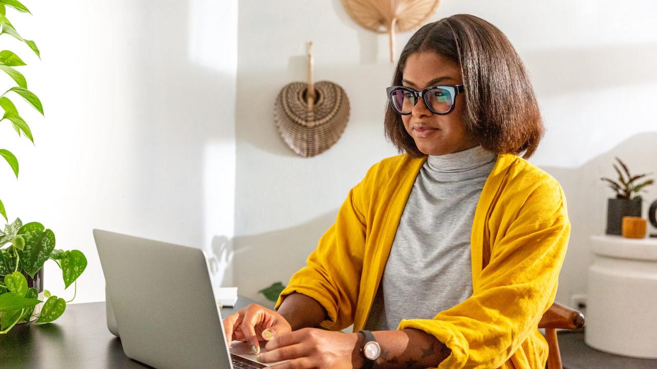 Black woman seated at a table and working on her laptop.