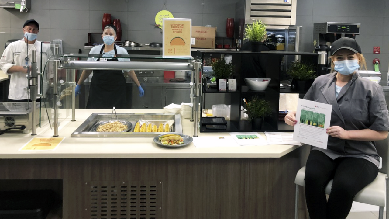 2 Blessing Hospital System masked employees stand behind a taco serving area in a cafeteria. An additional employee sits in front of the station, masked, holding up a document