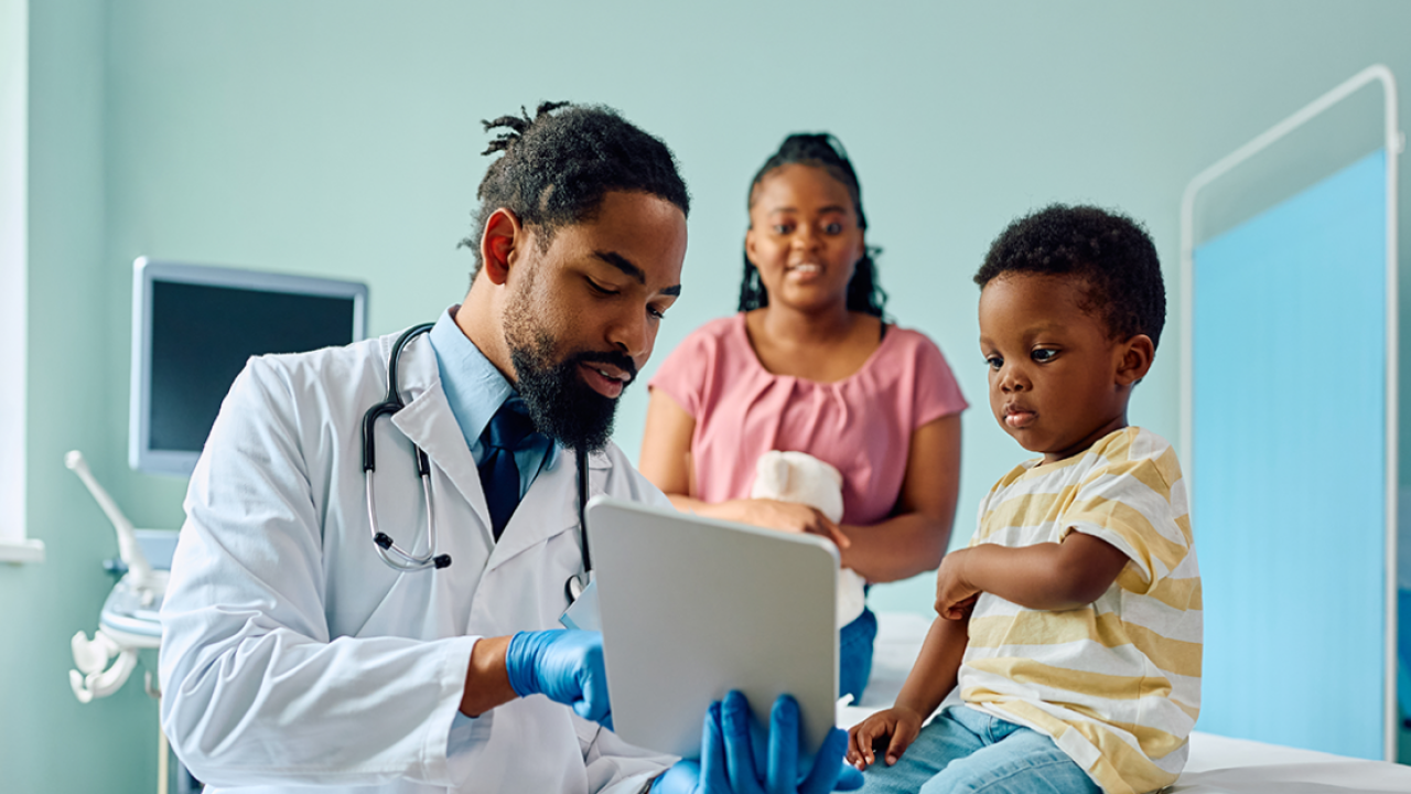 A doctor points to a tablet as a small child looks on. An adult smiling in the background
