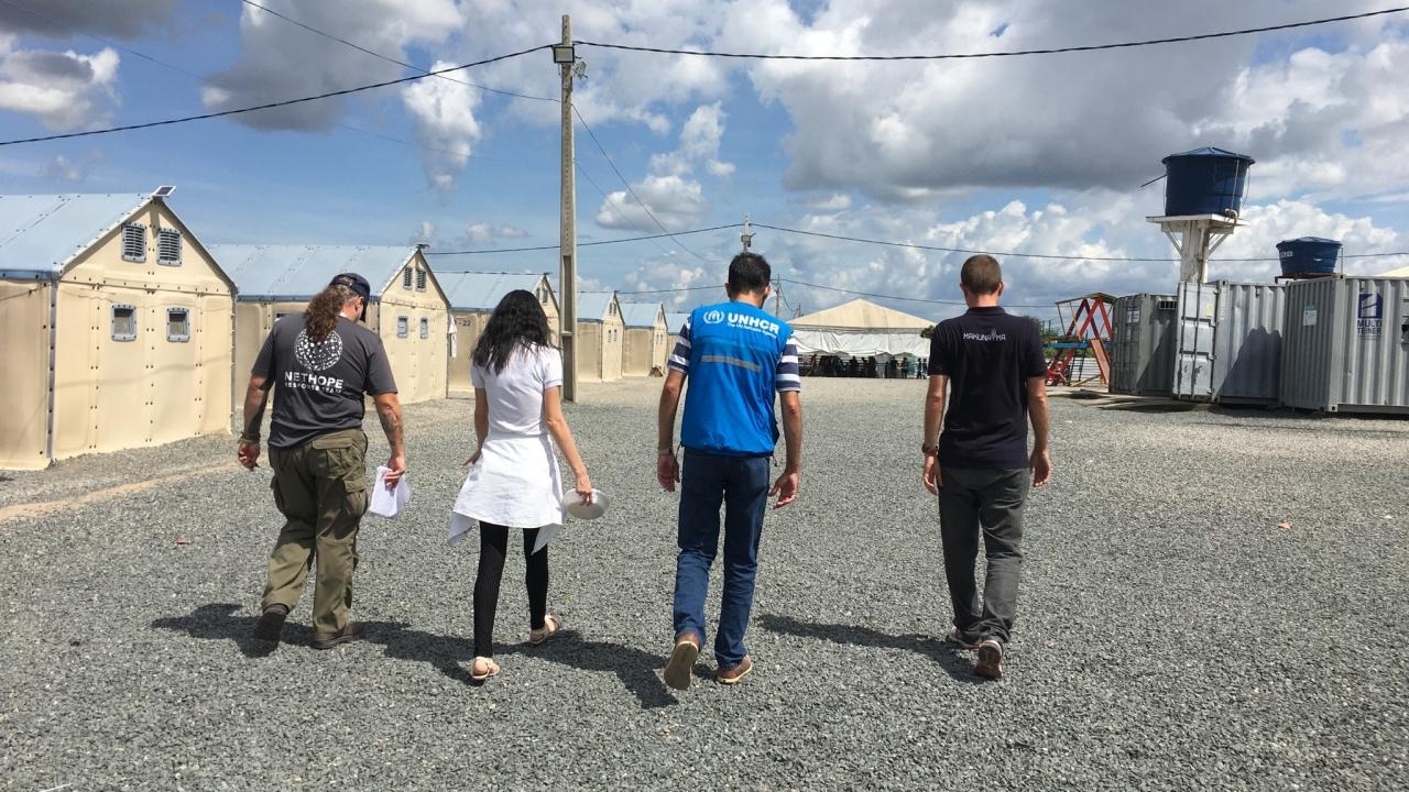 four people walking away from the camera on a gravel path. A row of Small brick buildings on their left and an open trailer on their right