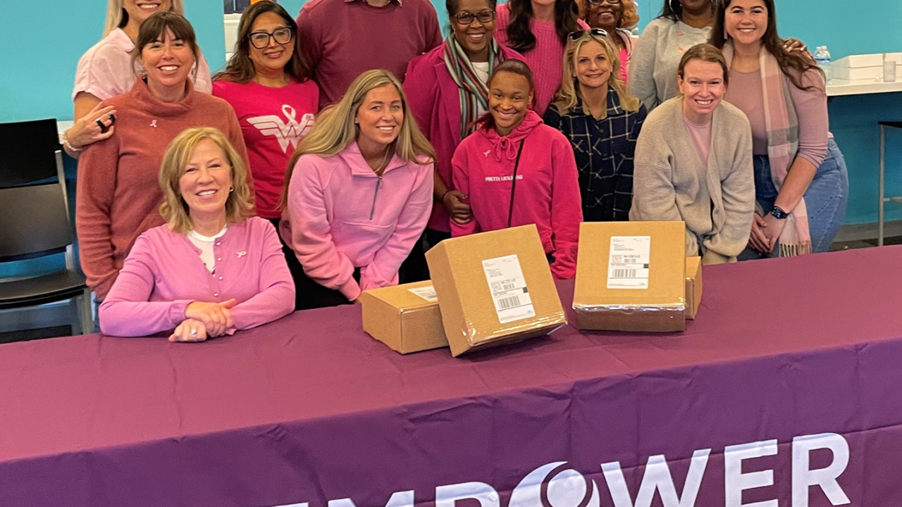 large group of volunteers dressed in pink pose behind a table that has an EMPOWER employee resource group tablecloth on it. On the table are Hope Kits.