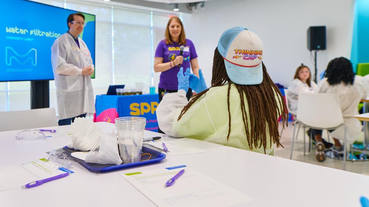 Students sit at a big table with paper and pencils while they talk with scientists at the head of a classroom.