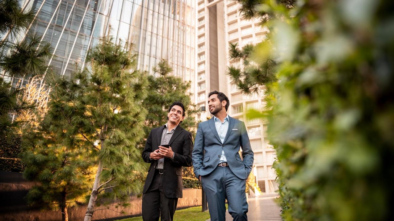 two people talk beside tall buildings and trees