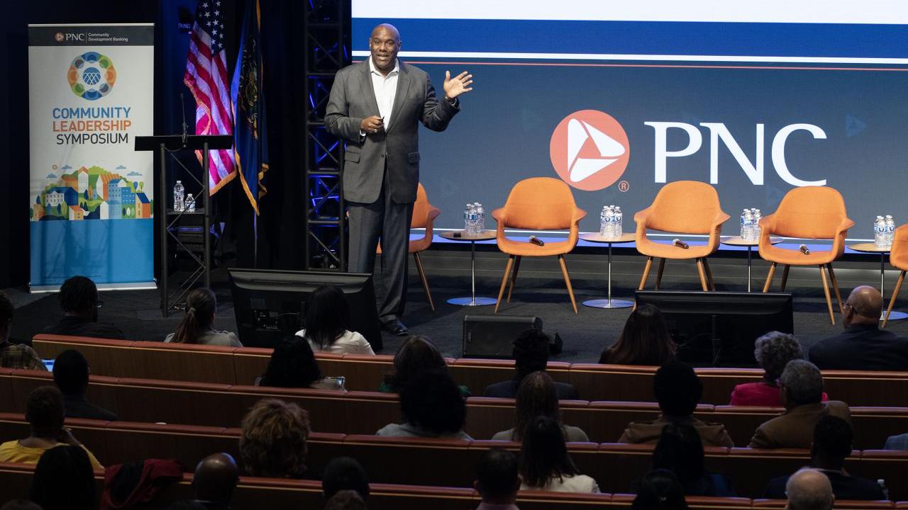 Richard Bynum standing on a stage in an auditorium of seated people. Chairs and PNC banner behind them.