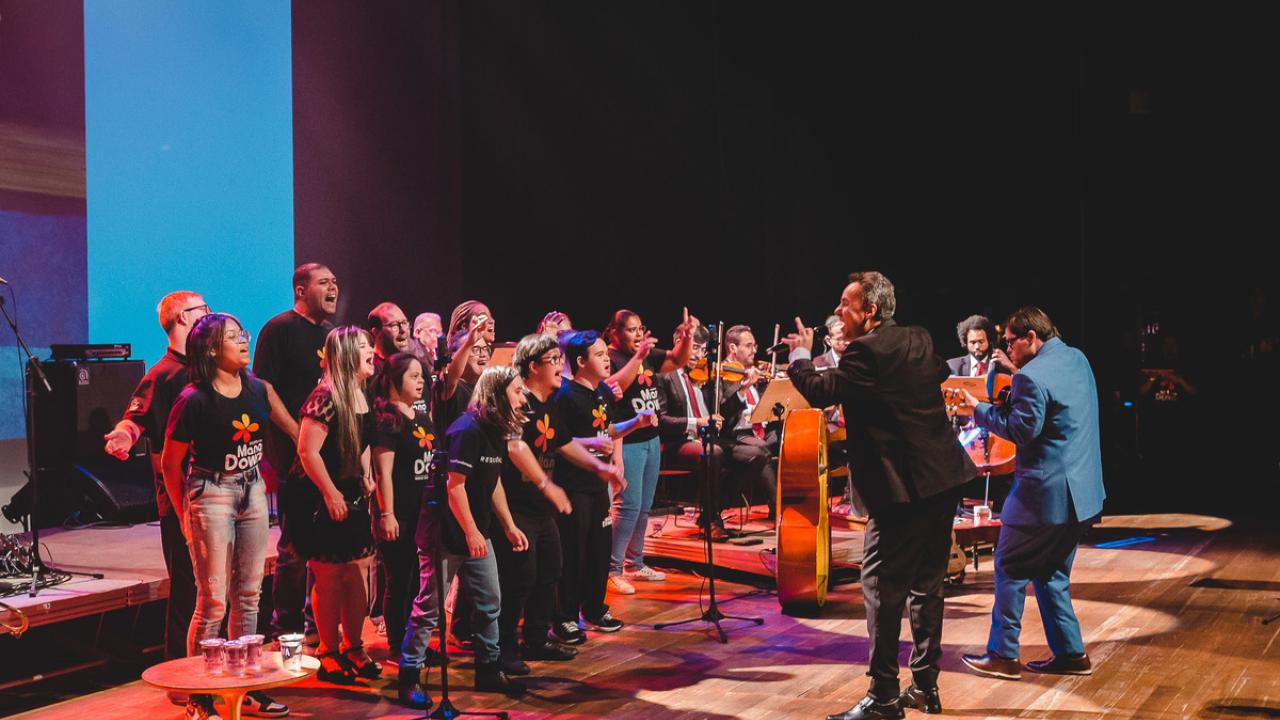 A group of students on stage with a small orchestra and conductor, all the students wearing "Mano Down" t-shirts.