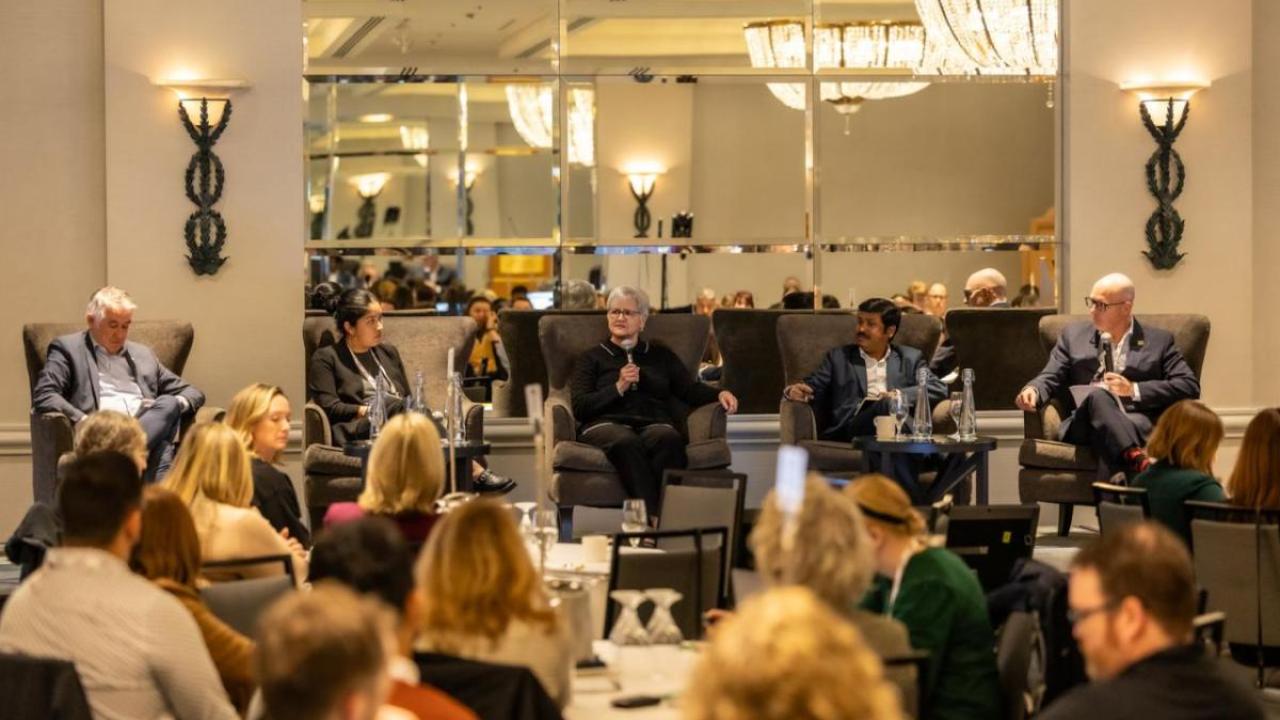A panel of five people speaking and answering questions at a conference event, seated on stage in front of an audience.