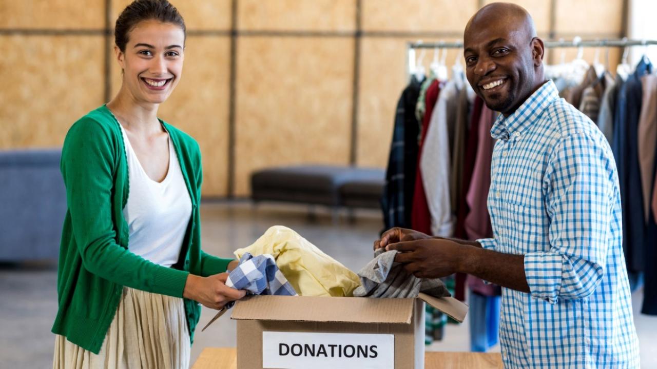 Two people holding clothing items in a "Donations" box. A rack of clothing hanging behind them.