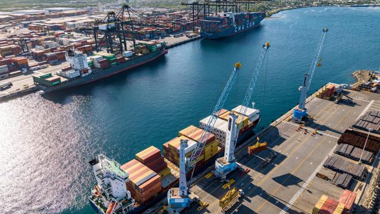 Aerial view of DP World port operations in the Caribbean, showing cargo vessels, cranes, and stacked shipping containers along the quay.