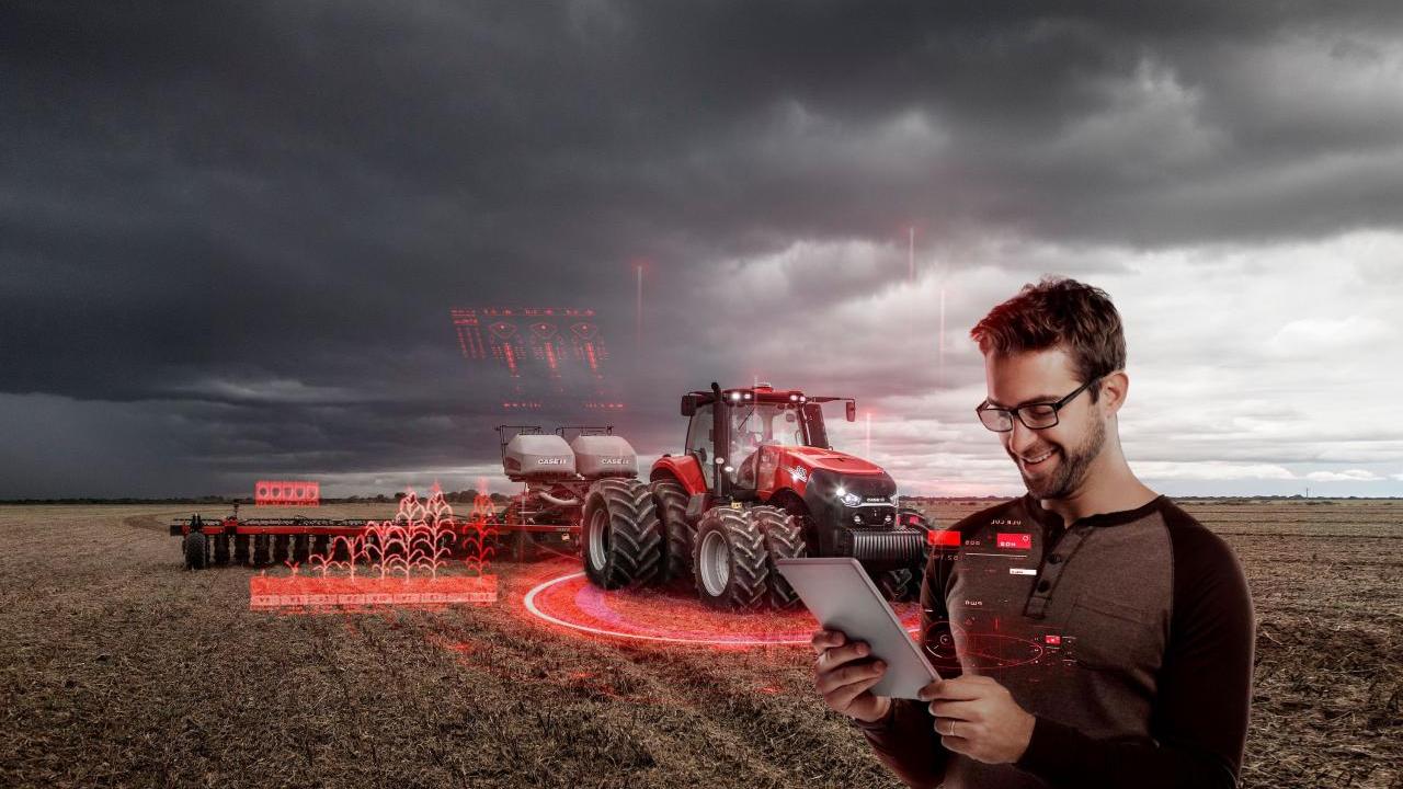 Red agricultural equipment in a crop field, with a person stood looking at a tablet PC