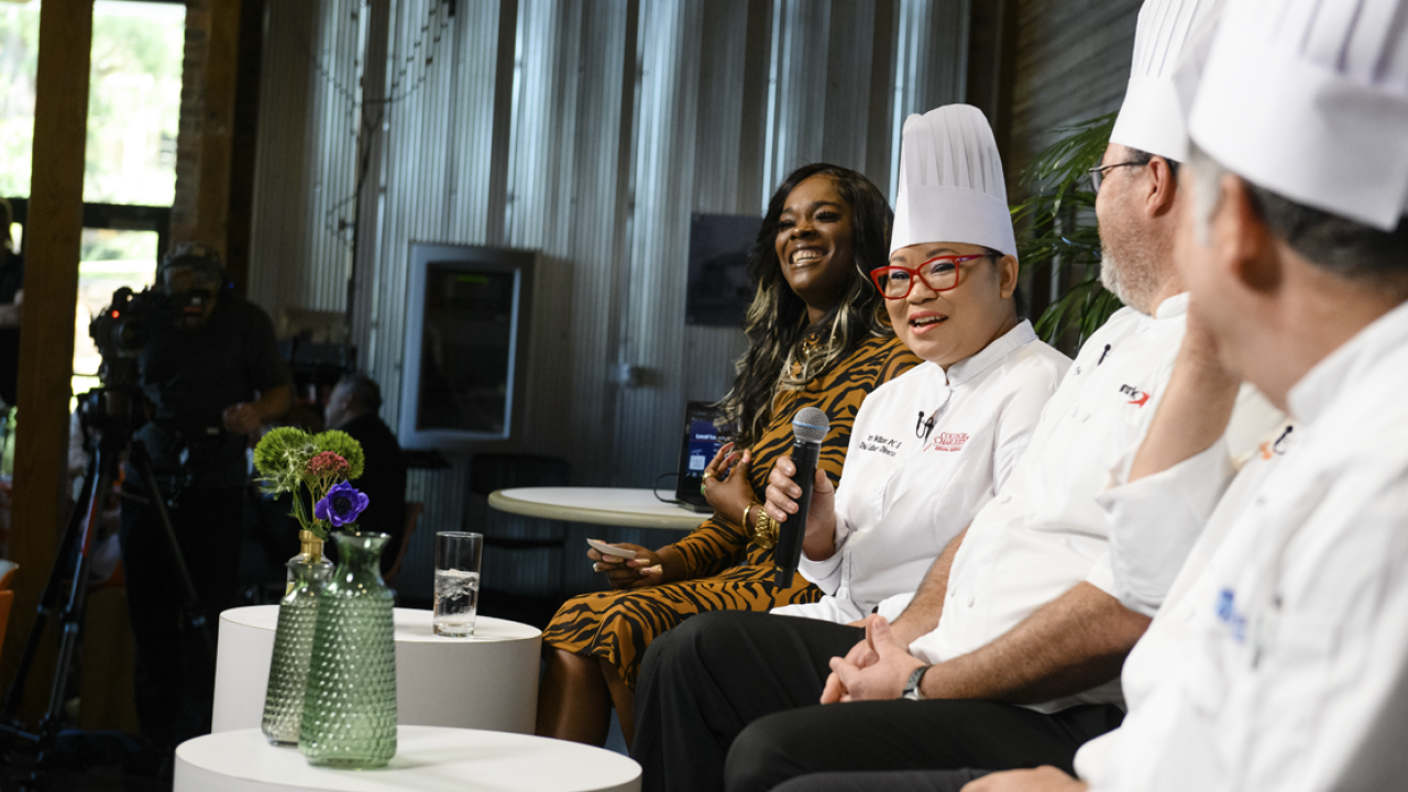 2 male chefs look at a female chef speaking as a part of a panel. The panel facilitator looks out into the audience.