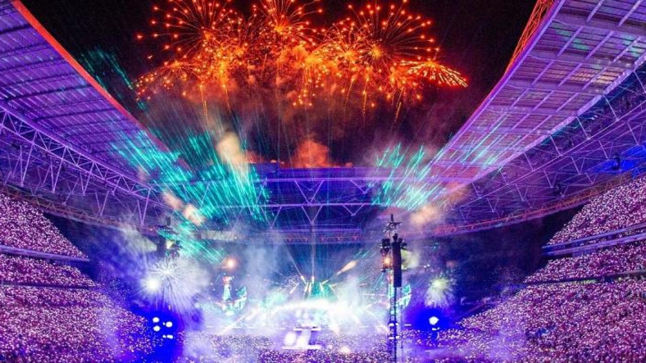 A huge stadium filled with spectators, a stage central is brightly lit, and orange fireworks fill the sky.