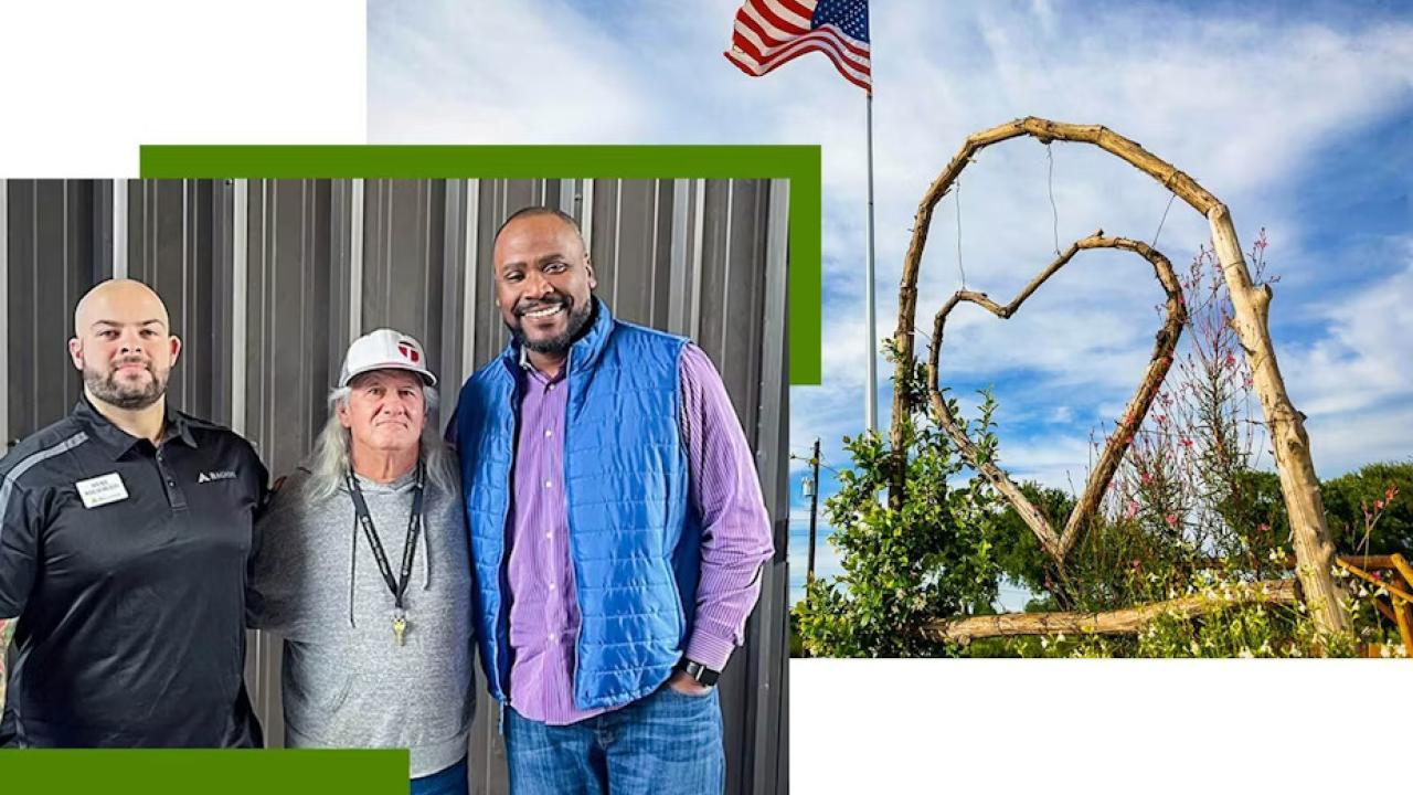 Three people with arms around each other. On the right A wooden heart hanging in an arch, an american flag on a pole next to it.
