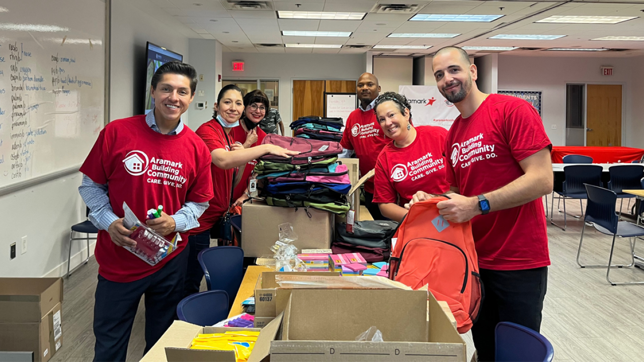 6 Aramark volunteers putting together backpacks, filling them up with assorted school supplies.