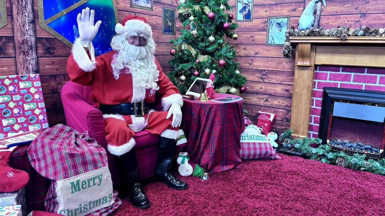 Santa waving at camera in front of Christmas tree and tablet