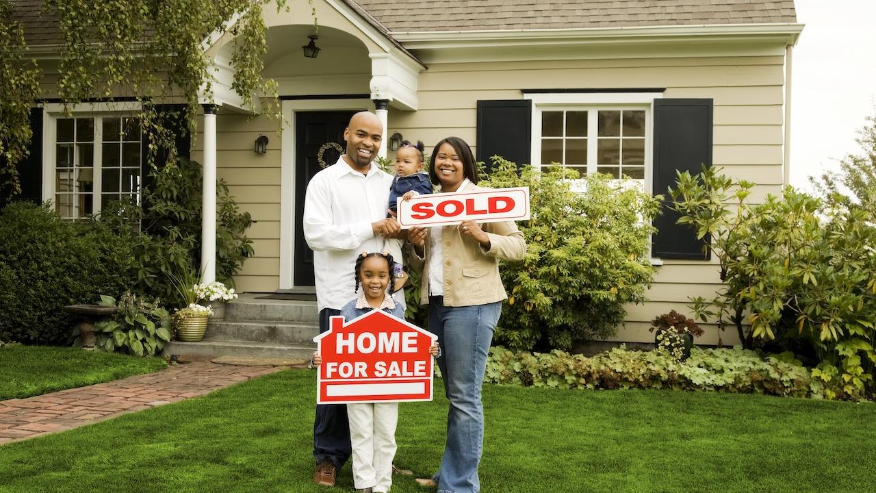 Young family of four in front of their new home holding a "sold" sign.