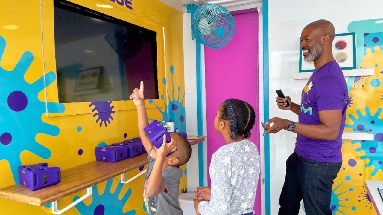 A smiling man stands next to two students who are engaging with interactive science materials. 