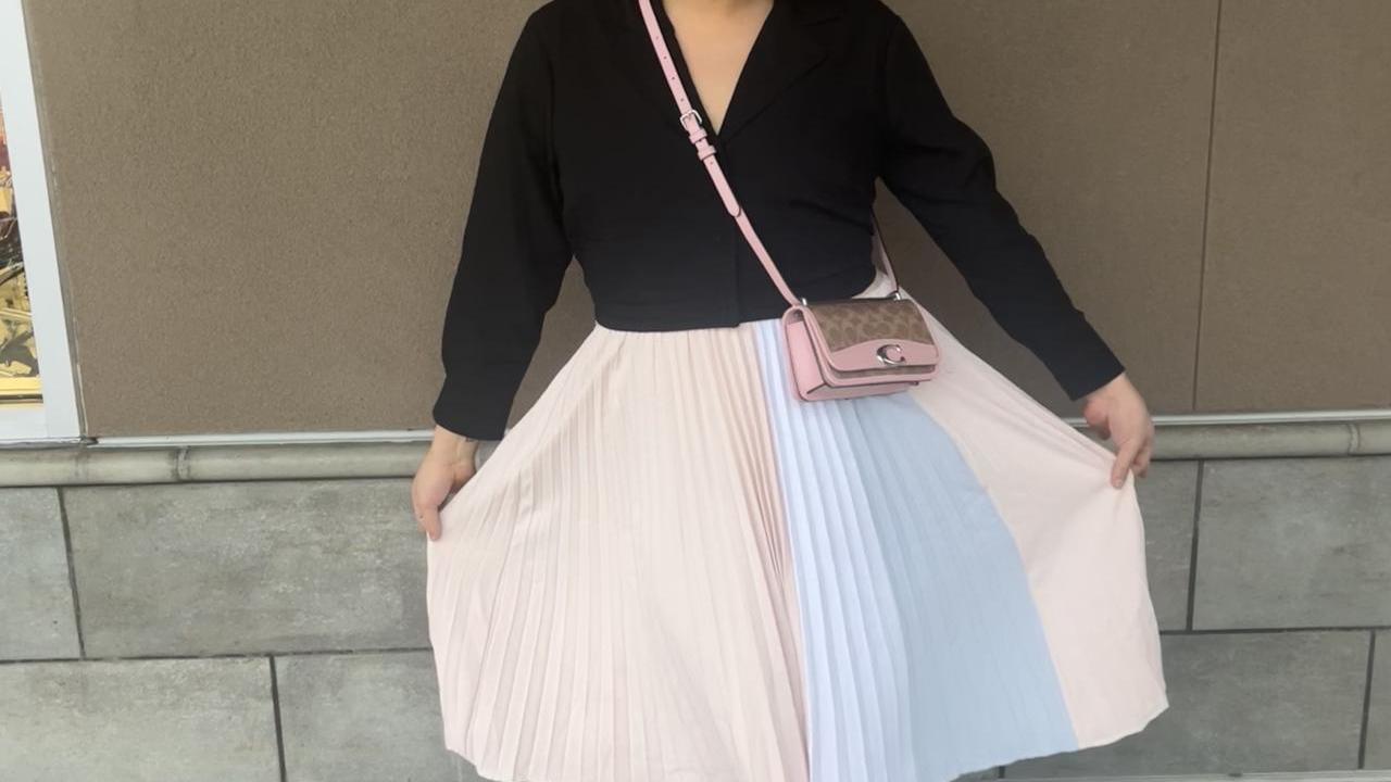 Curtlynn shows off her skirt, pleated pastel striped, strapy black heels and black v-neck top. She stands, smiling, in front of an exterior beige wall.
