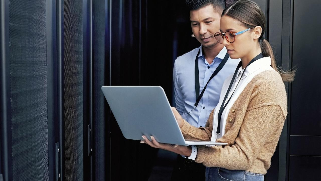 Two people in a server room and viewing a laptop.