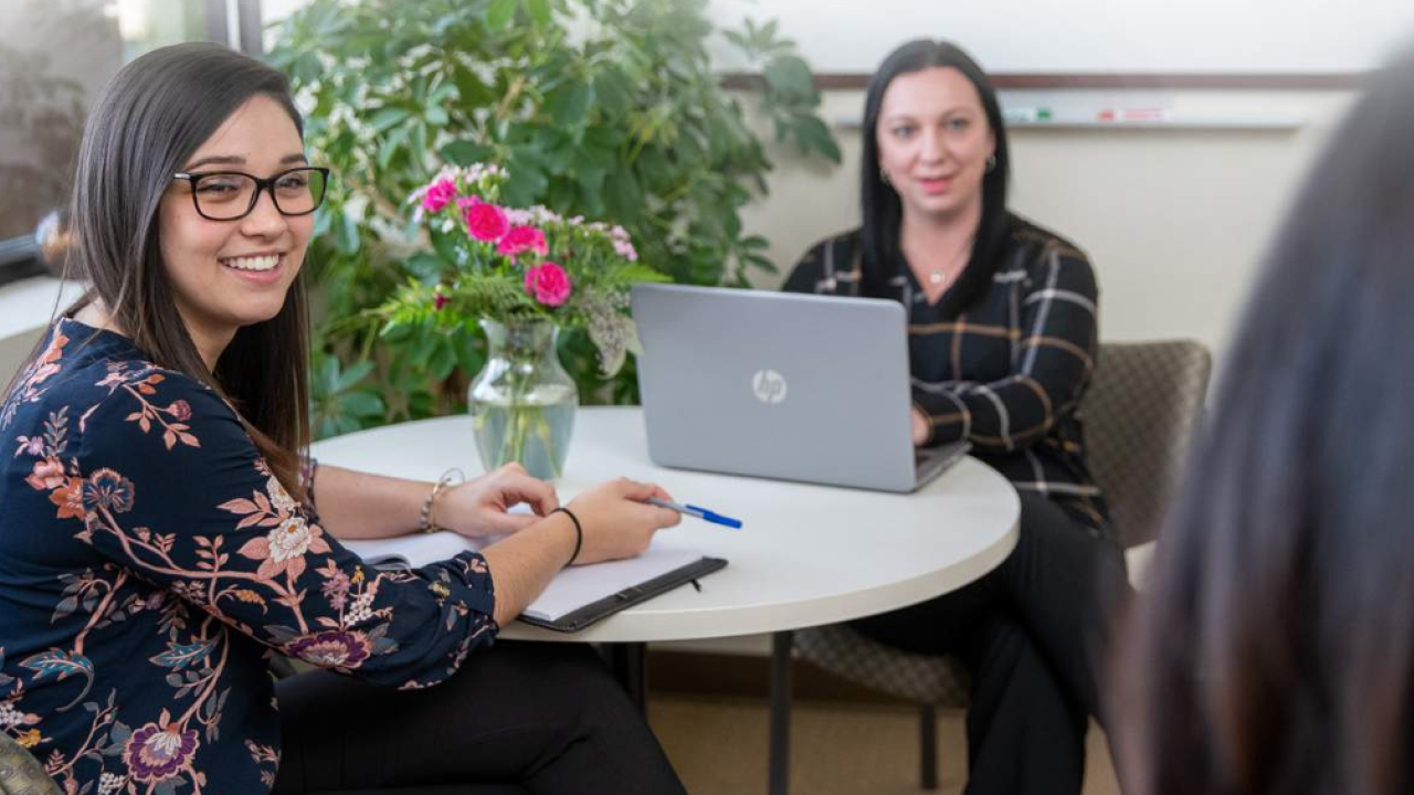 Two people sitting at a round table, both turned looking off screen. One has an open laptop.