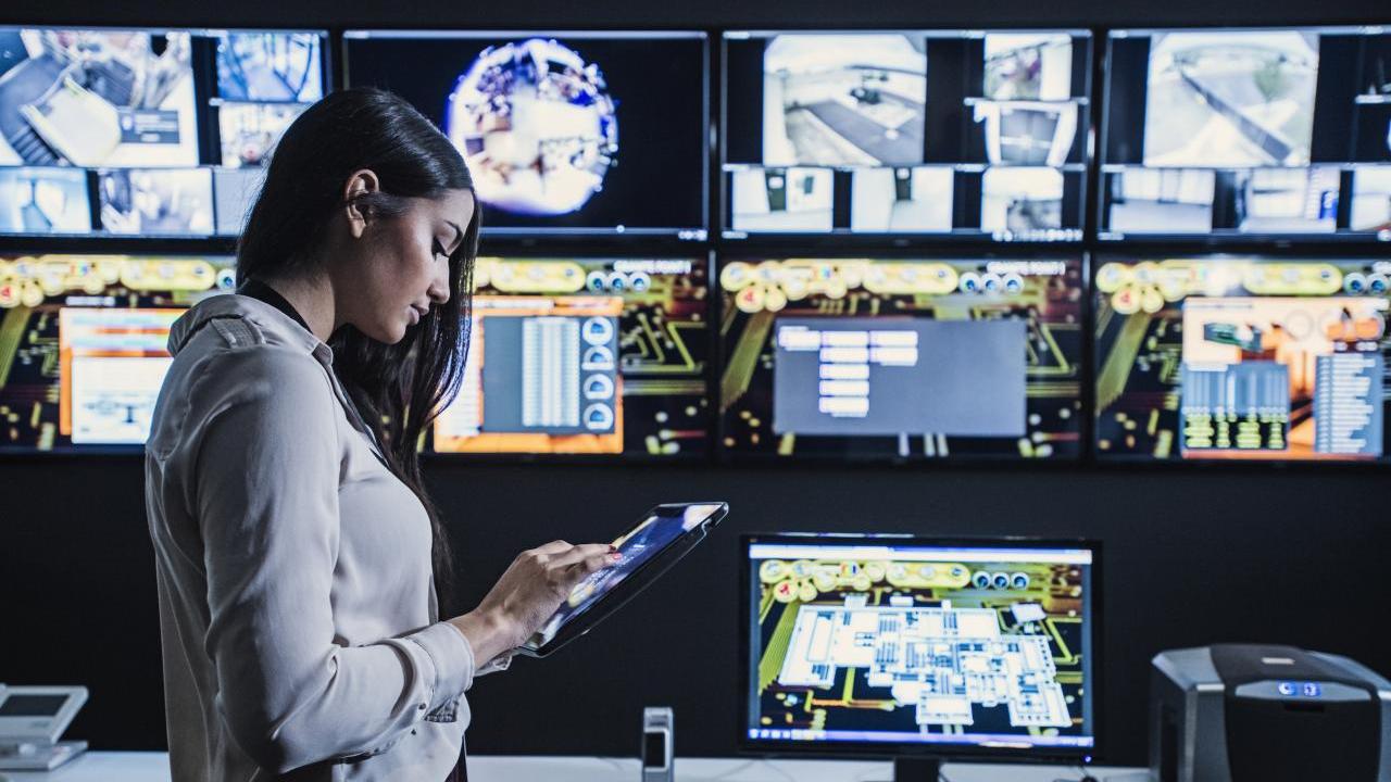 A person using a tablet in a room which has multiple screens on the wall and a computer on the desk