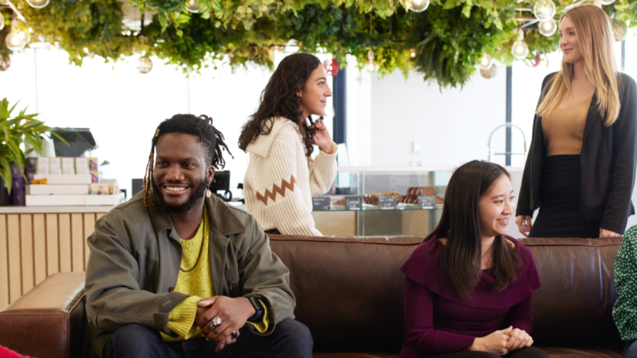 Group of people sitting inside on a couch or standing behind it, talking with each other. A cash register and bakery case behind them.