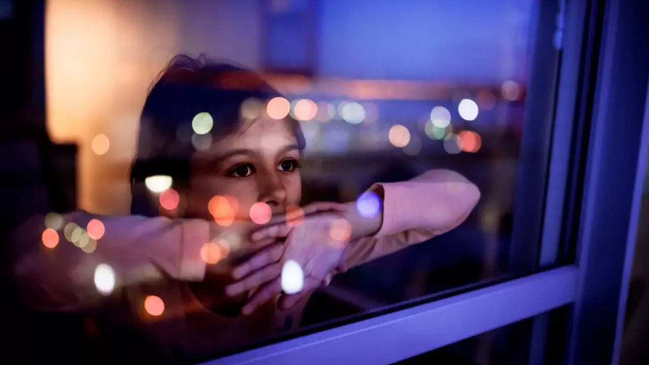 A child leant against a window with their arms folded, looking out