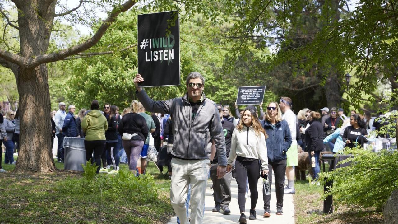A group walking down a sidewalk and gathered in a park holding a sign "I will listen".