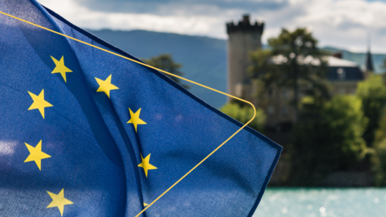 A flag and waterway, building, and scenery behind it.
