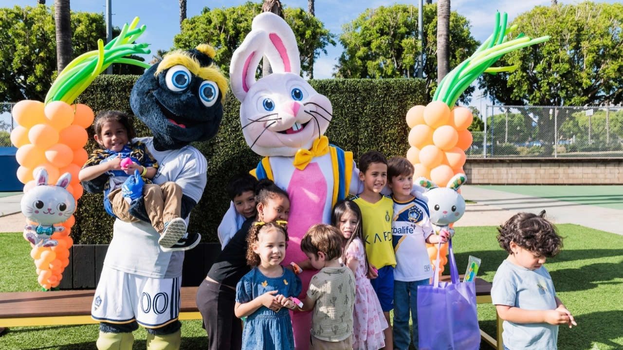 Children gather with two mascots on a turf field, holding gift bags and standing beneath balloon decorations.