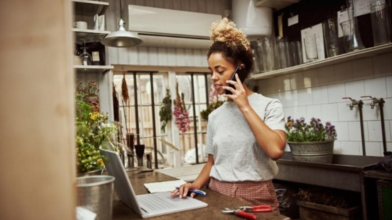 Young woman in a kitchen with her laptop open and speaking on a phone.