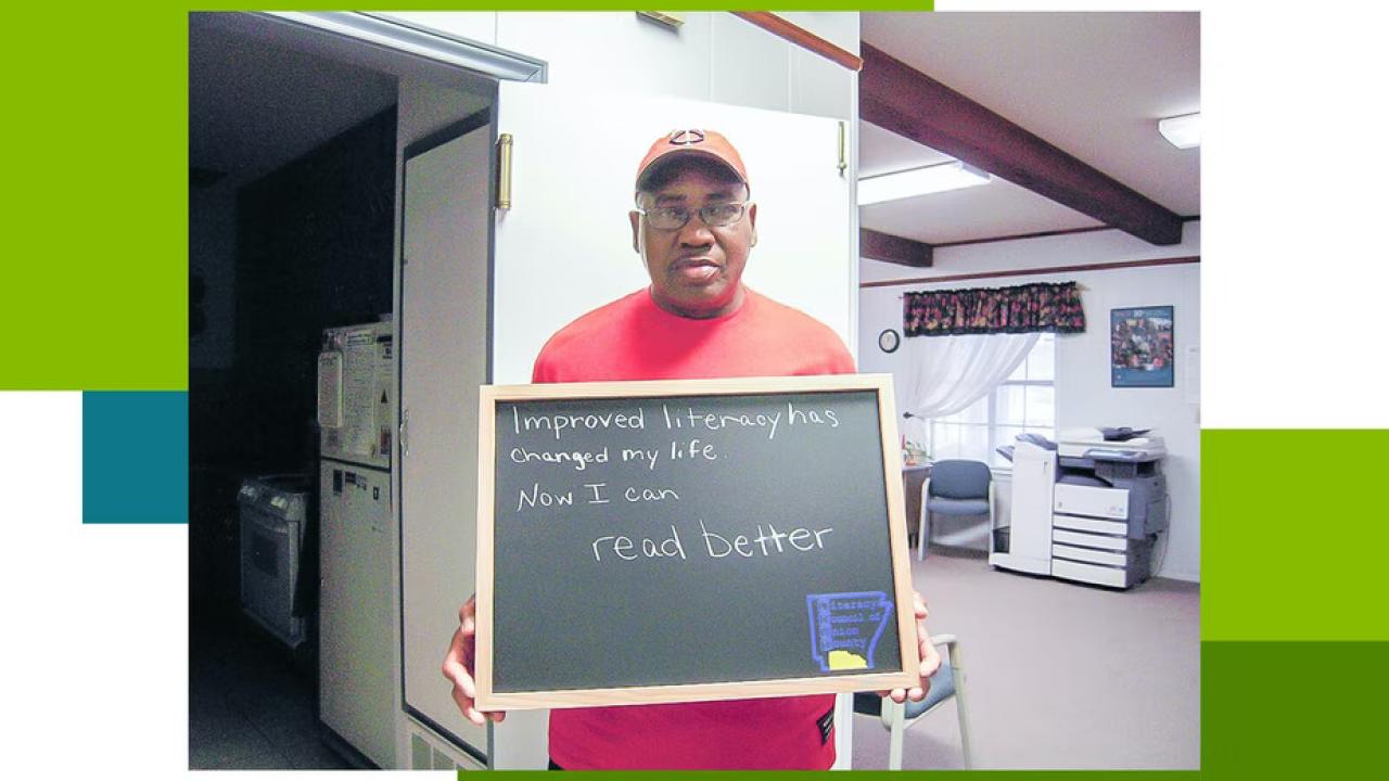 Person stands in a home setting holding a sign that reads "Improving literacy has changed my life. Now I can read better"