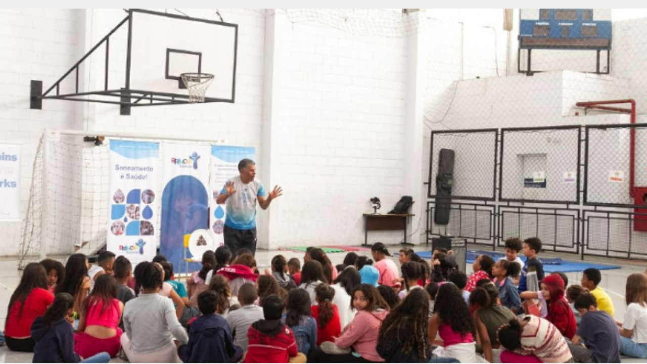 Person stood in a hall giving a presentation to students who are sat on the floor in front of them