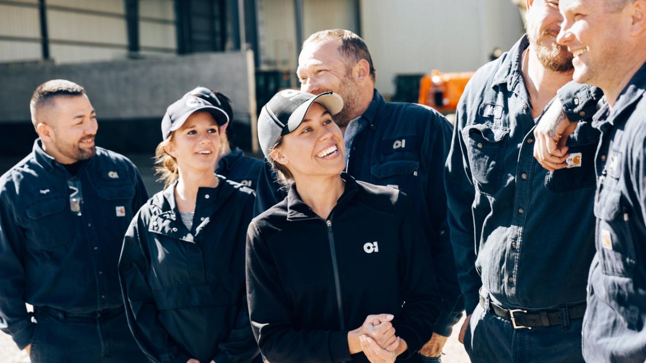 A group of people in dark work uniforms with OI logo, outside.