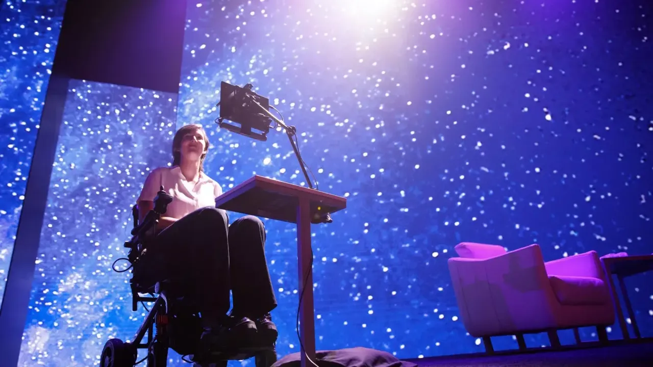 Erin on stage, a digital device in front of them and a colorful starry sky displayed behind.