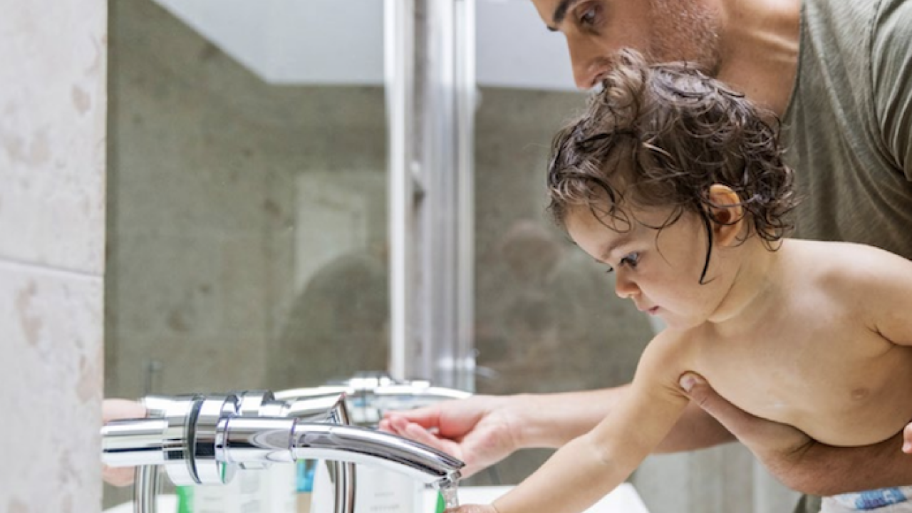 Man is holding a baby with his hand under a faucet.