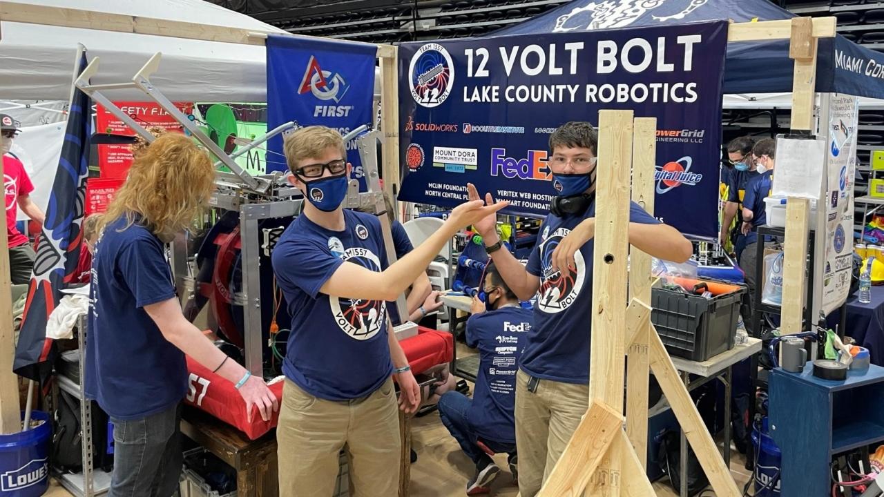 A group of students wearing the same "12 Volt Bolt" team t-shirts stand in front of their workspace
