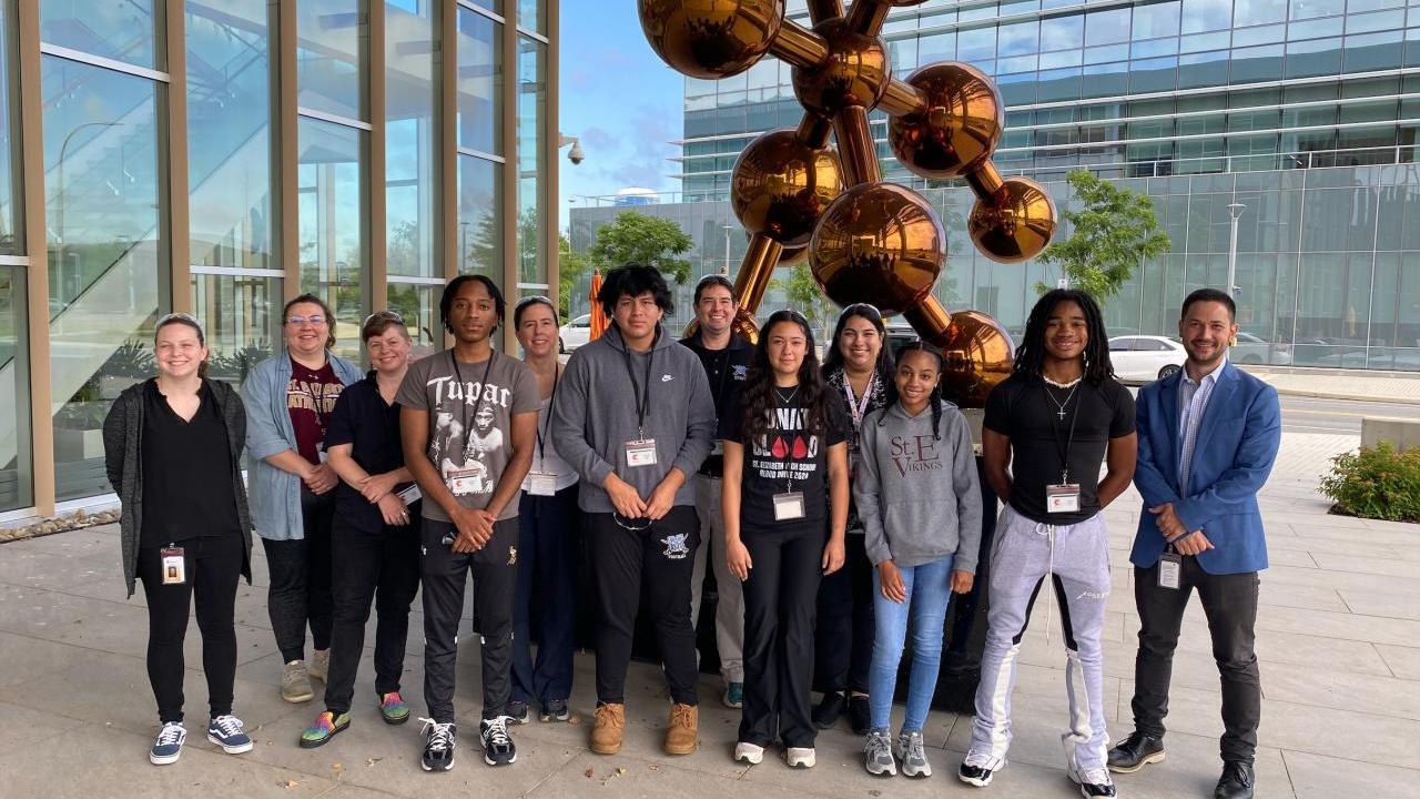 Students and adults posed next to a large molecule statue next to a large glass-sided building.