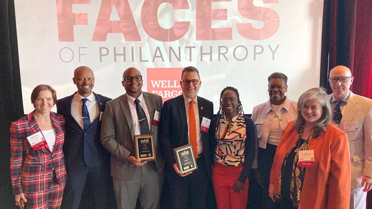 Group standing in front of a 'Faces of Philanthropy' banner. Two people are holding awards.