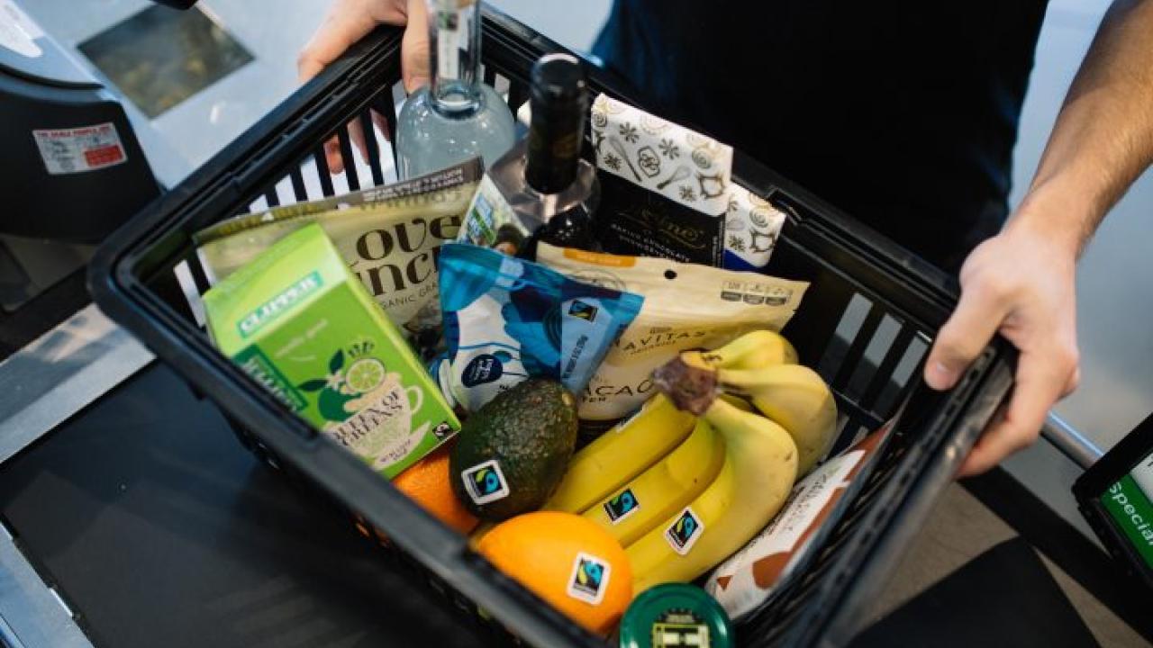 An assortment of Fairtrade certified products in a grocery basket, including bananas, avocado, and tea. 