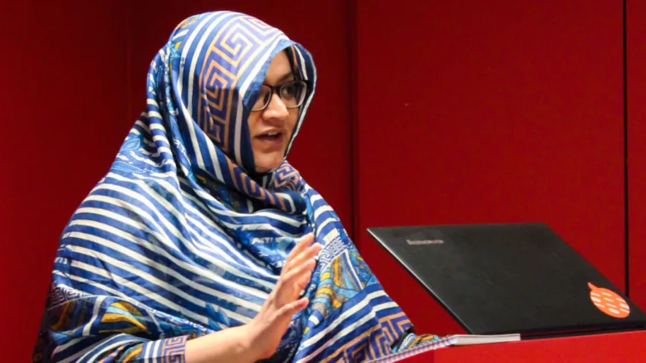 Faiza Yousuf standing at a podium with a laptop. Red walls behind her.