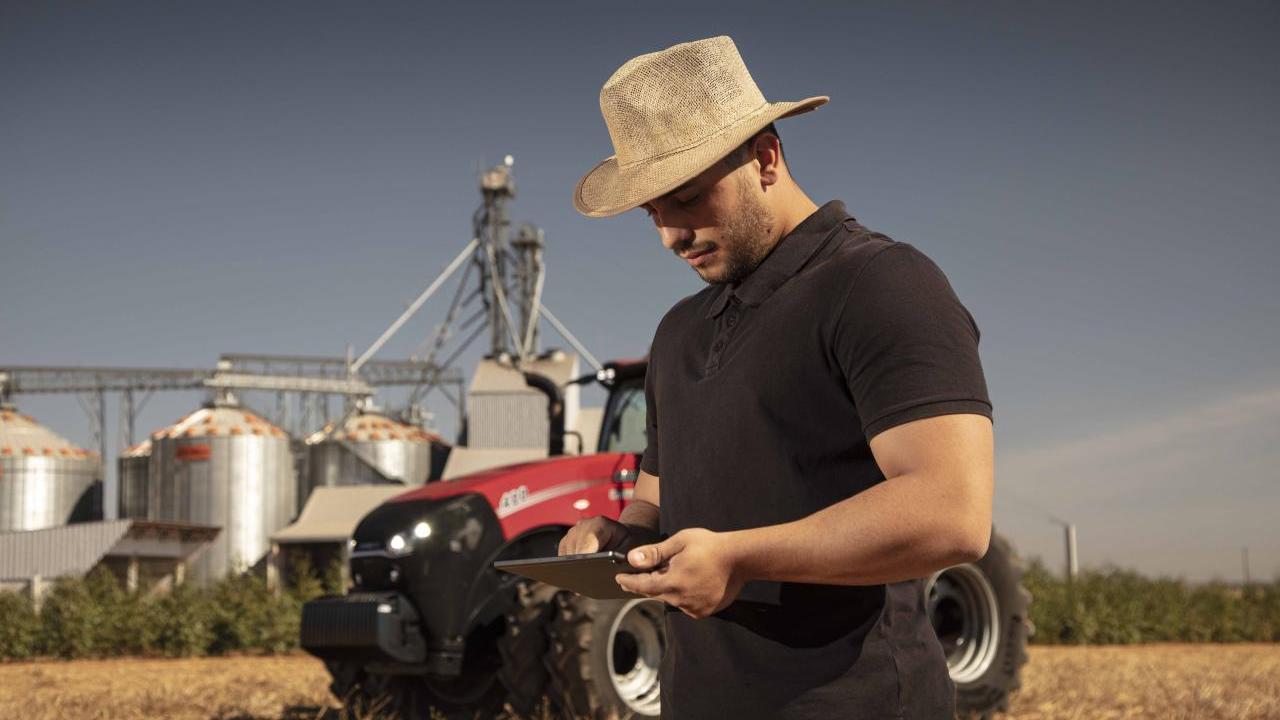 A farmer in a field looking at a tablet device. A red tractor and storage tanks behind them.