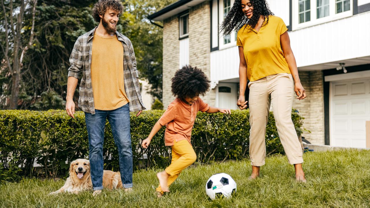Parents playing soccer with child and dog outside of house