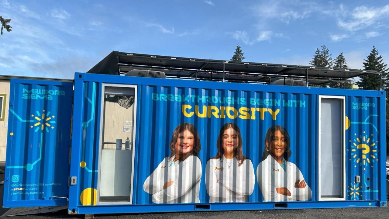 lays three The Curiosity Cube, a blue mobile science lab that displays three students in white lab coats and text that reads "Breakthroughs Begin With Curiosity"