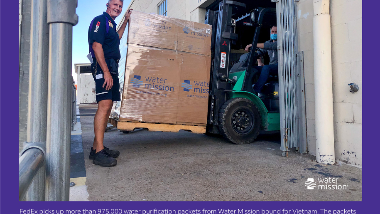 FedEx employee standing by forklift with large boxes labeled Water Mission. Caption reads "FedEx picks up more than 975,000 water purification packets from Water Mission bound for Vietnam. The packets will purify nearly 10 million liters of water.