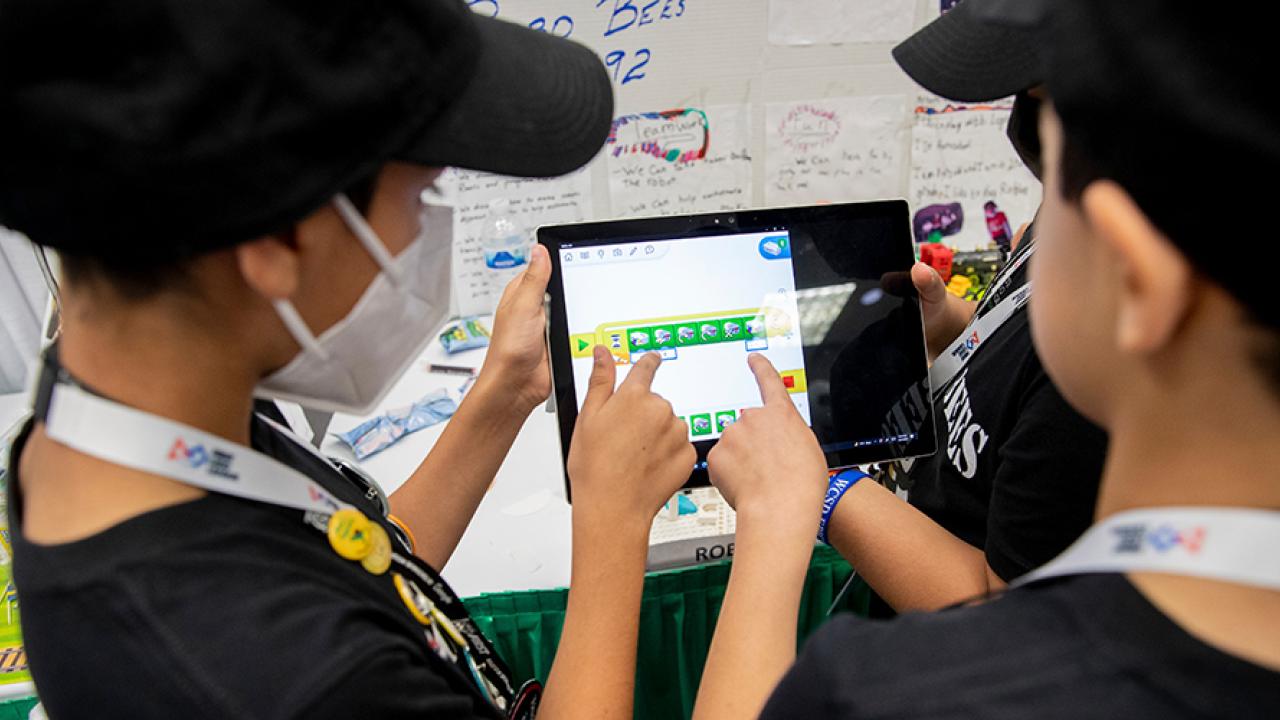 Children pointing at the same tablet, wearing matching lanyards and hats.