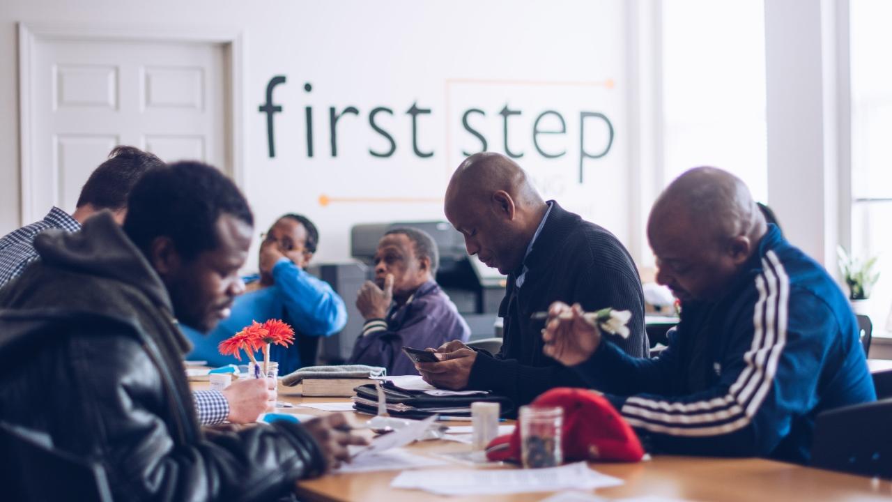 a group of people sitting at a table, filling out forms. "first step" banner in the background