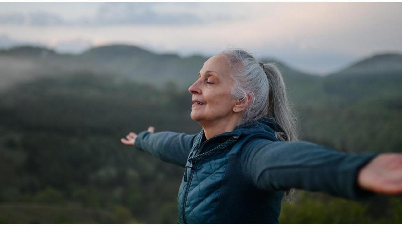 Person opens arms to sky with eyes closed with mountains behind them