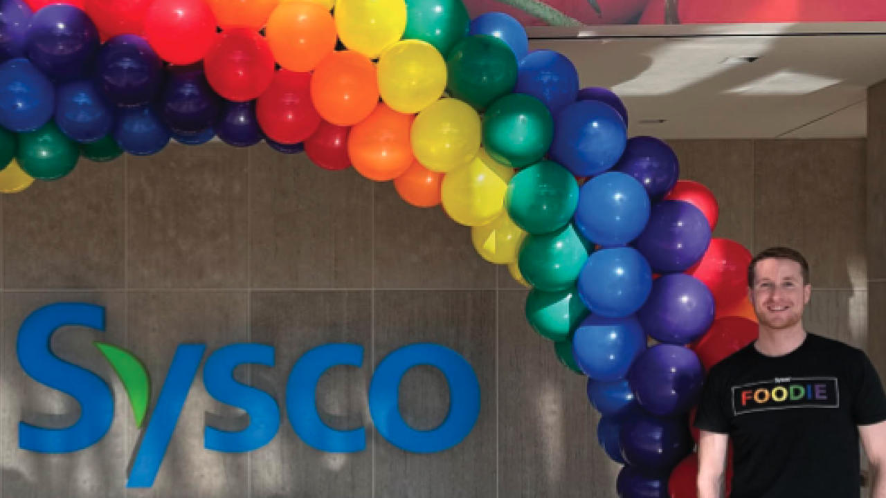 Person standing next to Sysco logo and rainbow of balloons. 