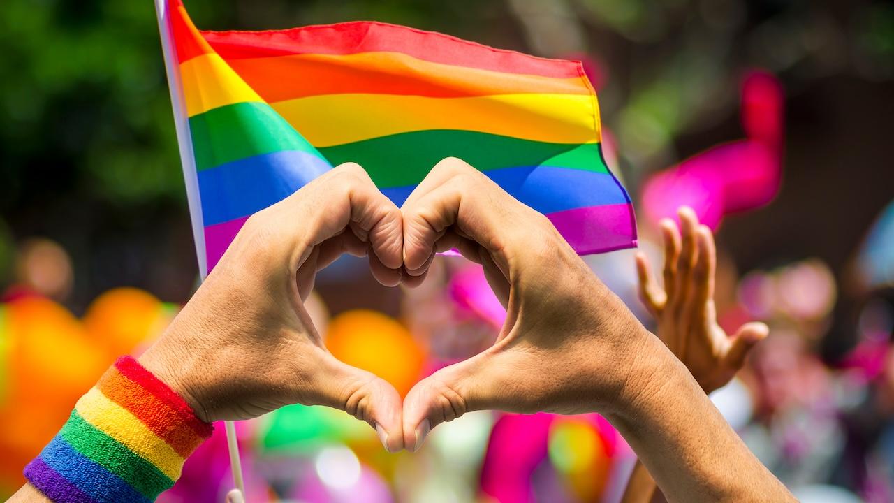Hand making a heart with a Gay Pride flag in the background.