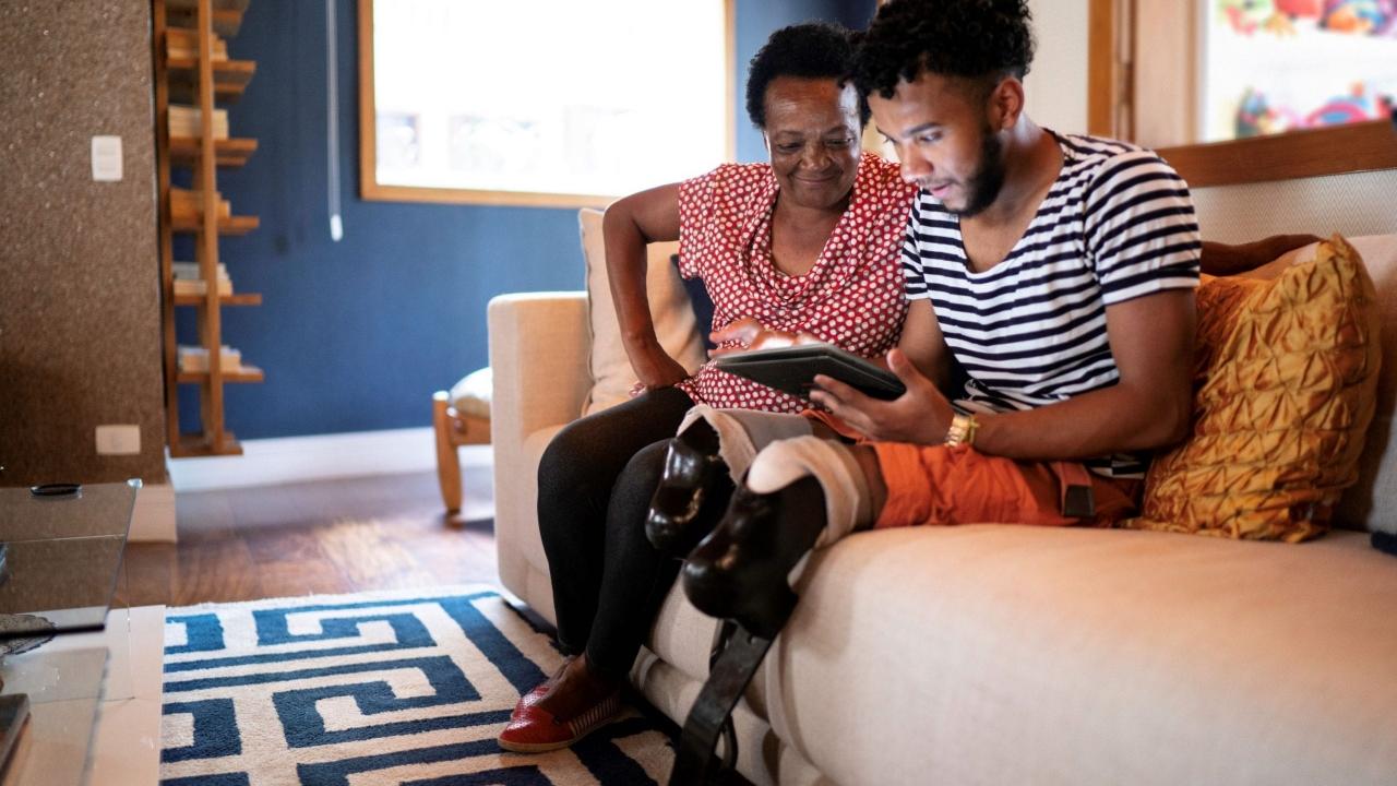 Photo of a woman sitting on a couch next to a man with prosthetic legs, both are looking at a tablet